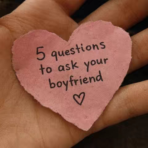 A hand holding a heart-shaped pink piece of paper; it's slightly textured and torn around the edges. On the paper, black handwriting spells out "5 questions to ask your boyfriend" in two lines, followed by a small heart doodle at the bottom.