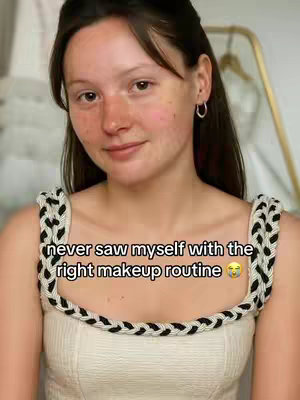 A young woman with fair skin and dark hair is the focal point, looking slightly to the left of the camera with a subtle smile. She is wearing a cream-colored top with decorative black and white straps. Text overlaying her face reads, 'never saw myself with the right makeup routine' followed by a smiley face emoji. The background appears to be a light-colored wall with a glimpse of what looks like a gold-colored clothing rack or mirror frame.