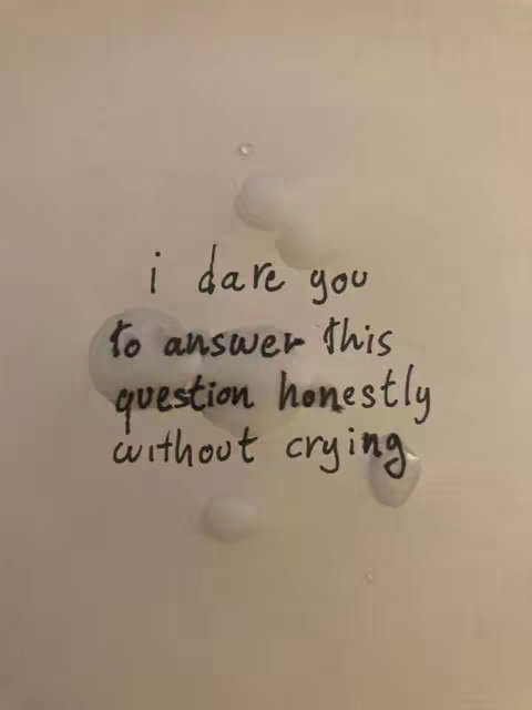 A close-up photo shows a white surface, likely a wall or a smooth, light-colored material, with handwritten text in black ink. The text reads: 'i dare you to answer this question honestly without crying'. Several irregularly shaped water droplets or condensation marks are scattered around the text, adding a tactile and potentially emotional context to the message.