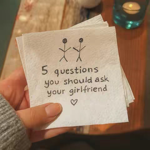 A hand holds a napkin with handwritten text and stick figure illustrations. The napkin is white with visible texture. The background includes a blurred tabletop and a partially visible candle and glass.