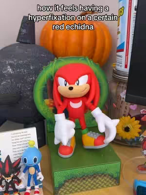 The image is a well-lit indoor shot featuring a Knuckles the Echidna figurine as the primary focus, standing atop a green platform. Behind the character, there's a medium-sized orange pumpkin. Several other objects, including a stone, and an unopened book, are arranged around Knuckles. The lighting appears natural, and the composition is such that the figurine is the clear focal point.