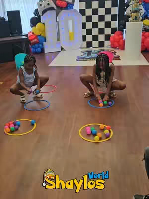 Two young girls are shown in a brightly lit room, squatting on the ground, each facing a colorful ball pit ball around a blue-colored hoop. The background features a '10' in large illuminated numbers, a black and white checkered backdrop, and balloons. The logo for "Shaylos World" is in the lower right corner.
