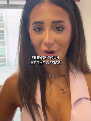 A woman with long dark hair looks directly at the camera, with a text overlay that reads 'FRIDGE TOUR AT THE OFFICE.' She wears a light pink top and has a necklace with a gold pendant. The background is slightly blurred, suggesting an office setting.