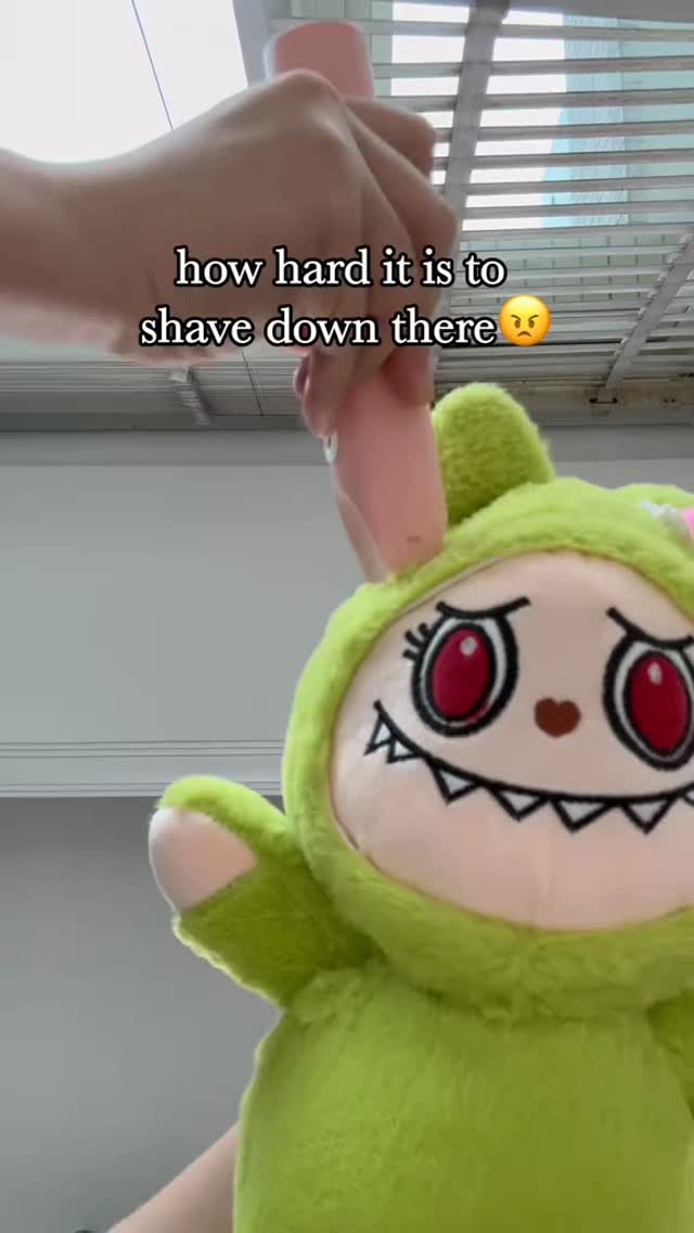 The image shows a plush toy shaped like a green monster with its face toward the camera. The hand of a person holding a pink electric groomer is seen. The image has text overlay.