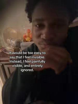 A dimly lit shot features a young black man looking towards the camera, slightly off-center. He is wearing a dark-colored garment. The background seems to be a warmly lit interior.