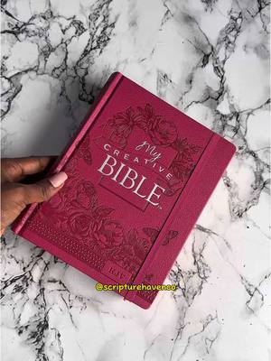 A close-up shot features a closed Bible held by a person's hand, with the book's cover prominently facing the camera. The cover is a deep, rich pink, with an embossed floral design and the words "My Creative Bible" in a clear, elegant font. The background is a marble surface with white and gray veining, and the composition is lit to highlight the texture and color contrast between the book and the background.