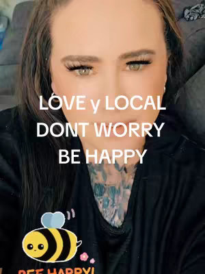 A close-up shot of a person's face with overlaid text. The person appears to be a woman with long, brown hair, wearing a black top. The text overlay reads "LOVE y LOCAL, DONT WORRY, BE HAPPY". A cartoon bee with a smiling face and the words "BEE HAPPY!" is visible on their top.