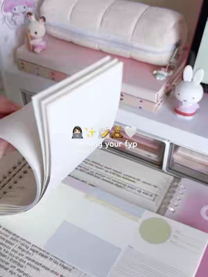 The cover image features a close-up shot of a person flipping through the pages of a notebook or planner. The background includes a desk with various stationery items, including a plush cushion, small figurines, and decorative boxes. The color scheme is soft and pastel, with pink and white tones dominating the scene. The overall composition is clean and organized, suggesting a study or workspace.
