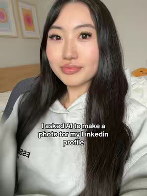 A selfie-style shot of a woman with long, dark hair and a gray hoodie. She's looking directly at the camera with a slight smile. Text overlayed says 'I asked AI to make a photo for my Linkedin profile'. The background includes two framed pictures and a bed.