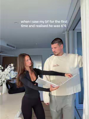 A young woman in a black outfit is holding a white document, positioned in front of a taller man in a cream-colored sweater and gray pants. They appear to be indoors, with a mirror and some white curtains visible in the background.