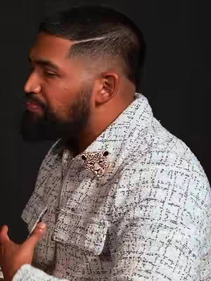 A man in a textured white and gray jacket with a decorative pin is captured in a medium shot, turned slightly to his left. His hair is neatly styled, with a groomed beard and mustache. The lighting highlights his features, suggesting a setting of moderate contrast. Behind him, a dark background creates a contrast.