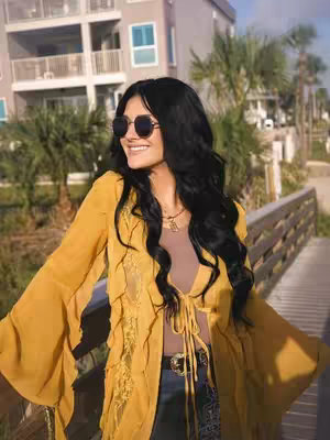 A woman wearing black sunglasses is standing on a wooden bridge. She is wearing a yellow jacket over a brown top and jeans, and she is smiling, with buildings and palm trees in the background. The color palette is dominated by warm tones.