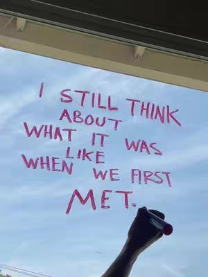 A hand holding a red marker writes text on a window, with the phrase "I STILL THINK ABOUT WHAT IT WAS LIKE WHEN WE FIRST MET." The sky is visible in the background.