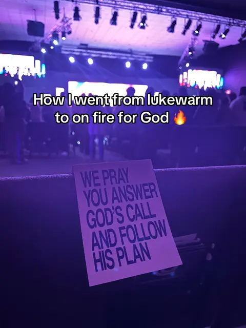 A photograph taken inside what appears to be a church or large assembly hall. The perspective is from a seated position, looking towards the stage. A sign reading 'WE PRAY YOU ANSWER GOD'S CALL AND FOLLOW HIS PLAN' is prominently placed in the foreground, in front of the seats. The background includes a stage or platform, with people visible in the background and overhead stage lighting. The overall lighting is dim, with a purple hue.