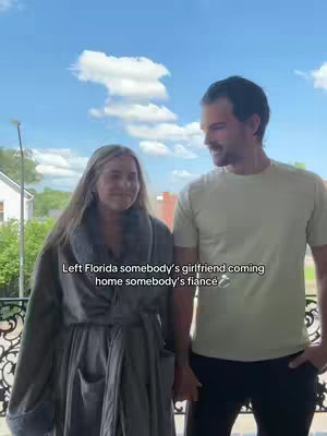 A couple stands close together on a balcony with a blue sky and clouds in the background. The woman on the left is wearing a gray robe, and the man is wearing a light-colored t-shirt. Both are smiling. Text overlay at the bottom reads, 'Left Florida somebody's girlfriend coming home somebody's fiancé'