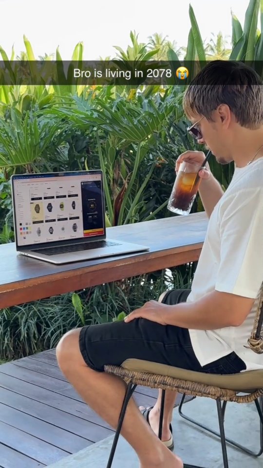 A man is sitting at a wooden table outside, holding a glass of a dark drink with a straw, with a laptop open in front of him. Behind him are lush green tropical plants. The text overlay on the image says: "Bro is living in 2078" and a crying face emoji is present.