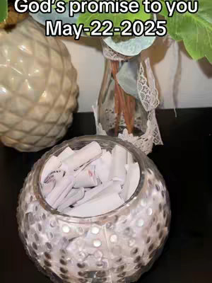 The image showcases a glass container with small white paper rolls inside, suggesting rolled-up notes or messages. In the background are blurred elements including a vase with flowers and greenery. Overlayed at the top: text over a blurred background stating "God's promise to you May-22-2025". The overall composition uses a soft color palette.