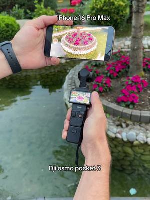 A person is holding an iPhone 16 Pro Max (top) and a DJI Osmo Pocket 3 (bottom). The person is standing near a body of water with greenery and flowers in the background. The iPhone is displaying a scene from the camera it is filming. The DJI Osmo Pocket 3 is also displaying a scene from its camera. The background includes water, green bushes, pink flowers, and concrete.
