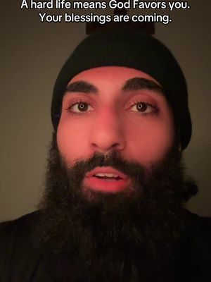 Close-up shot featuring a man with a long black beard and a black beanie. He's in front of a background with dim lighting. The text overlay reads: 'A hard life means God Favors you. Your blessings are coming.'