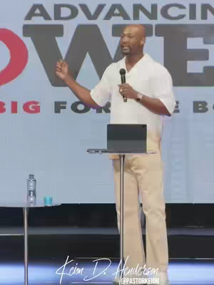 The image shows a man in a white shirt and light-colored pants speaking into a microphone on a stage. Behind him is a large screen with the words "Advancing Power" and "Big For The Box." He has a serious expression and is gesturing with his right hand. The stage has a podium with a laptop on it and a small table with a bottle of water and a blue cup. The image is well-lit and appears to be taken during a sermon or public speaking event.