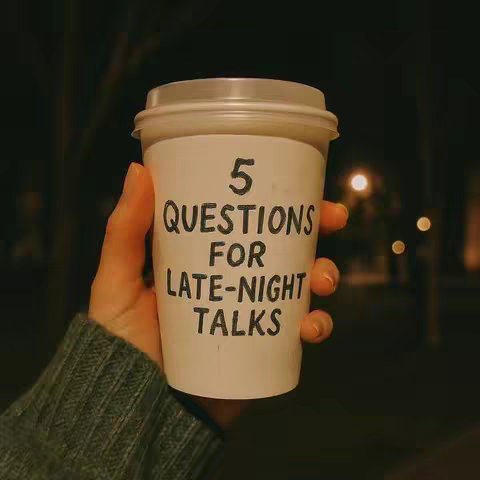A hand holds a white paper coffee cup with a clear plastic lid against a blurry background, likely at night. The cup has text on it.