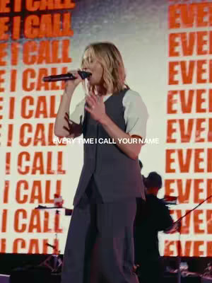A person is singing with a microphone on stage, a background of vertical text with the words "I CALL" repeating, with overlaid text "EVERY TIME I CALL YOUR NAME".