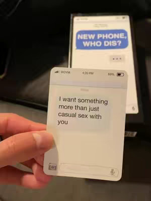 The image shows a smartphone displaying a text message conversation on a white background. The conversation shows the text bubble reads "NEW PHONE, WHO DIS?" On top of this, the hand is holding a white card printed to look like an iphone displaying another text message, that reads: "I want something more than just casual sex with you". The whole image is shot in a way to emphasize the messages.