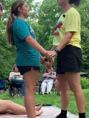 Two young women holding hands and looking at each other, standing in a backyard setting. Other people are in the background sitting in chairs.