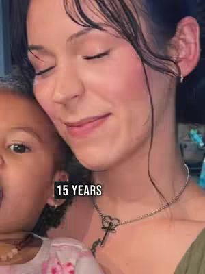 A young woman and a small child are in a close embrace. The woman's eyes are closed, and she is smiling. The child is looking at the camera with a slight smile. The image has a soft, warm tone.