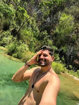 A man stands in a shallow pool of clear, turquoise water, with lush green foliage and a rocky cliff face in the background. He is smiling with his right hand on his forehead.