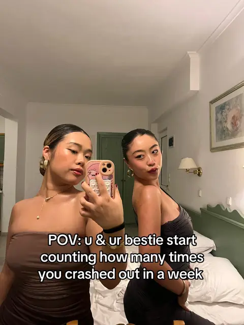 Two young women are posing for a selfie indoors, likely in a hotel room. The woman on the left is holding the phone-camera, while the woman on the right is leaning back. Both are dressed in evening wear. The background features a bed, a framed picture, and a wall with a neutral color profile.