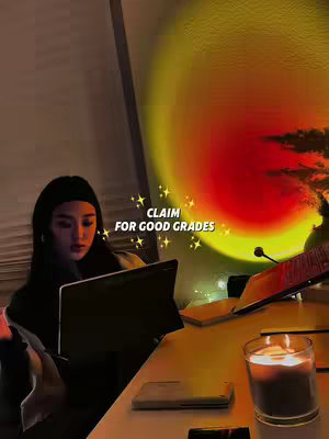 A woman is seated at a desk, working on a laptop. The scene is dimly lit by a warm, orange-toned light, with a candle and other objects on the desk. Text overlays read 'CLAIM FOR GOOD GRADES' with star sparkles and other details.