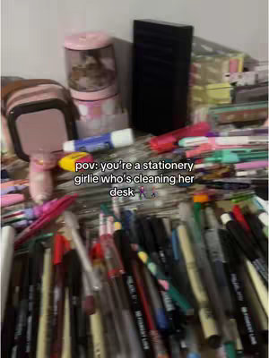 A cluttered desk covered with pens, markers, a pencil sharpener, a glue stick, and a pink bag. The image gives a first-person POV of a student's messy desk.
