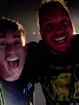 Two men are centered, close, and smiling widely, with their mouths open in the cover image. The man on the right has a dark shirt with yellow graphics, and the man on the left wears a black shirt over a chain. The background is dark and indistinct.