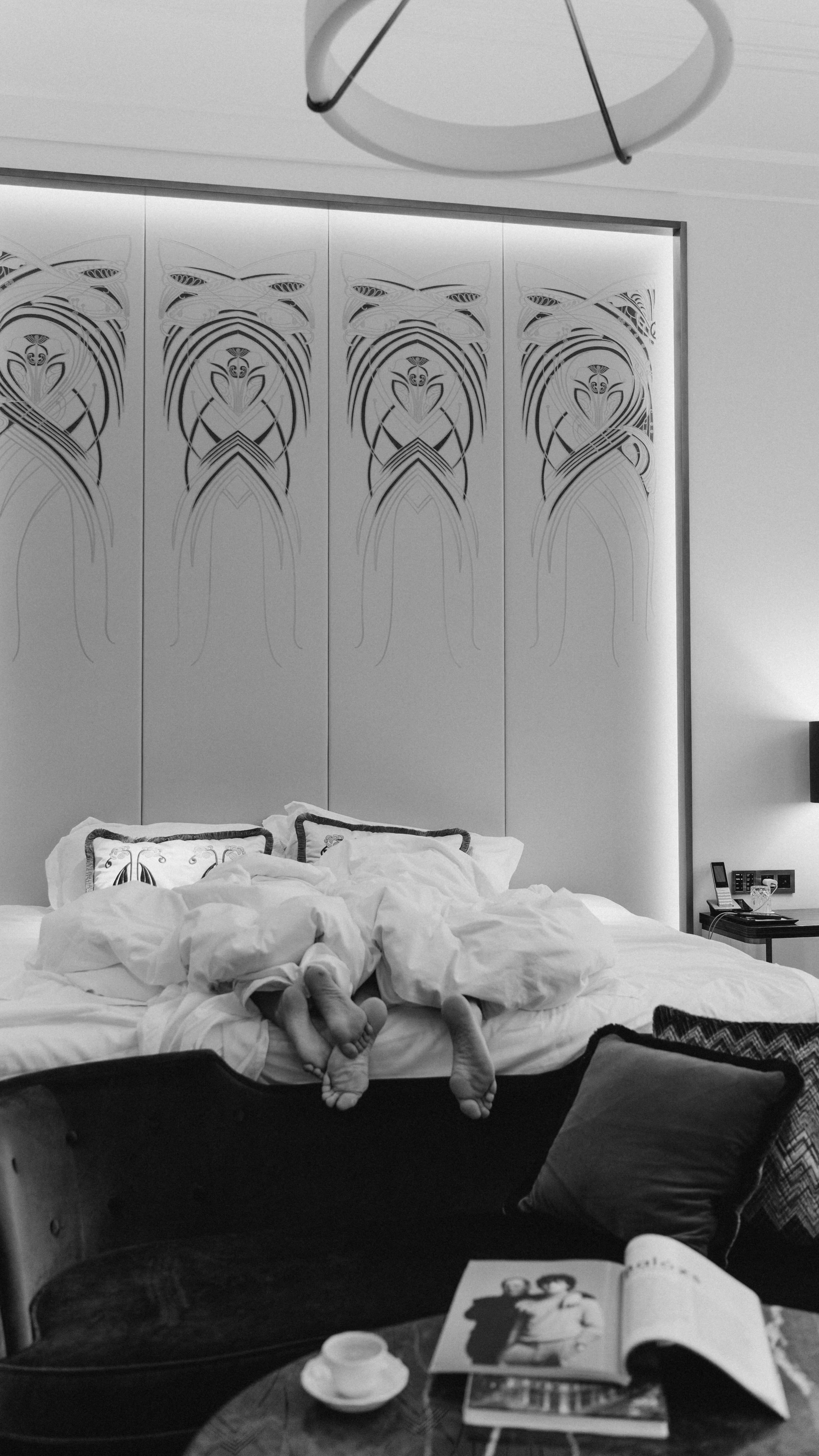 Black and white image of a hotel room bed with two people's feet sticking out from under the covers at the bottom of the frame. The bed has white bedding and decorative pillows. In the background is a patterned wall. A table with items on it is visible to the right and a sofa along the left.