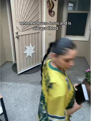 A woman with a black ponytail, walking past a beige front door with a decorative design. She holds a black book in her right hand. The image appears to be taken outside.