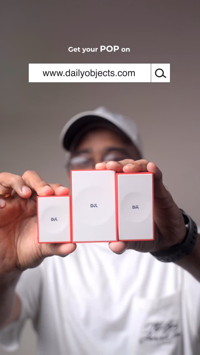 A person is holding three white and red rectangular power adapters. Above the person is the text 'Get your POP on' with a search bar displaying 'www.dailyobjects.com'. The items are being held in front of a neutral background with the person wearing a white shirt and a cap.