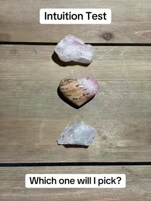 The image shows three crystals on a wooden surface. The top and bottom crystals are roughly shaped and white or translucent. The middle crystal is heart-shaped, with a mix of brown and pink hues. Text overlays are present at the top and bottom, with the top text reading 'Intuition Test' and the bottom text reading 'Which one will I pick?'