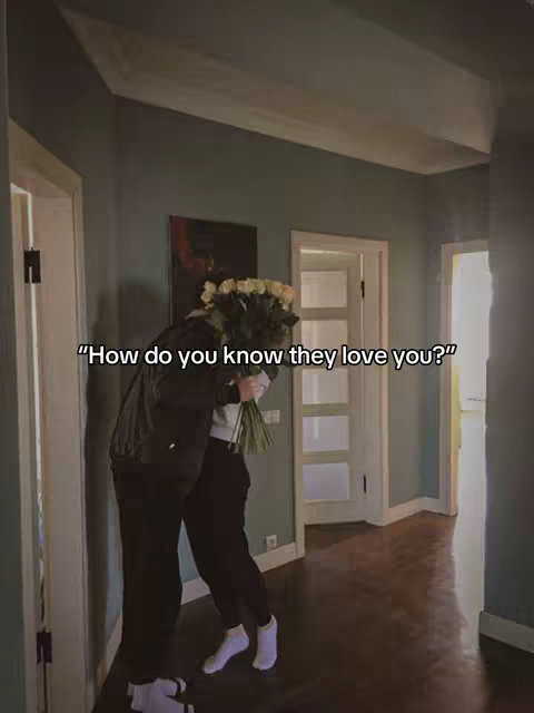 An interior shot shows a hallway with two figures embracing. One figure holds a bouquet of yellow roses, partially obscuring their face. The hallway has walls painted in muted tones, with doors on either side. Text overlayed on the image poses a question about love.