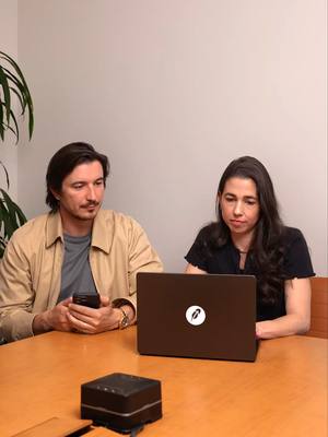 A photo of a man and a woman sitting at a table, facing a laptop. The man is on the left, wearing a light tan jacket over a grey t-shirt. He's holding a phone and has a watch on his wrist. The woman is on the right, wearing a black shirt, and looking at the laptop screen. The laptop is closed.