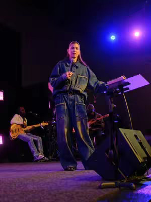 The image features a woman standing on a stage, holding a microphone, with a music band playing in the background. The setting appears to be a church or a similar venue, with stage lighting highlighting the woman and the band. The woman is wearing a denim jumpsuit. The composition includes a lectern or music stand, and the overall atmosphere suggests a performance or a devotional session.