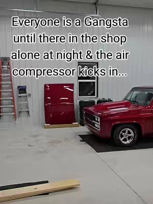 The image shows a well-lit shop interior with a red pickup truck parked on the right side. The image includes a ladder, a red panel, a small window, and tires.