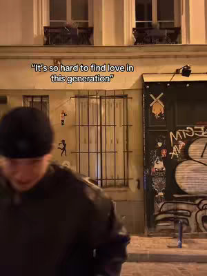A person in a black beanie and jacket is shown in the foreground against the backdrop of a street scene at night. The background features buildings with architectural details such as windows and a painted door. The image is illuminated by streetlights.