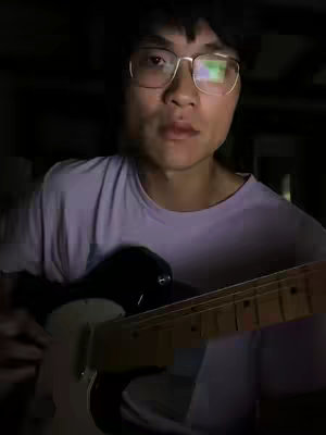 A man with glasses is holding a guitar in a dimly lit room. The lighting is focused on his face. The background is dark.