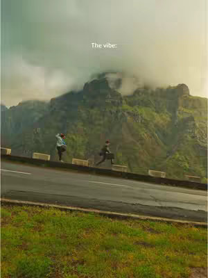 A scenic shot showcasing a mountain range in the background with a road and grass in the foreground. Two individuals are pictured walking on the side of the road.