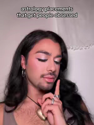 A person with long dark hair is styled, with a subtle makeup look and accessories. The person is angled towards the camera, gesturing with a finger, and appears to be talking. The background is simple.