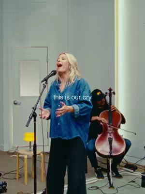 A live music performance scene. A woman is center stage, singing into a microphone with her arms outstretched, while a cellist plays behind her. The stage has basic lighting, and the background suggests a studio setting.
