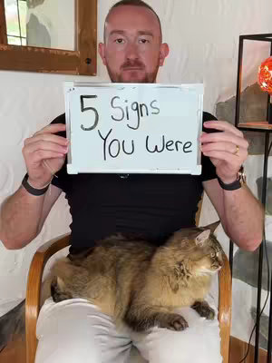 A man with red hair sits in a wooden chair, holding a small whiteboard that reads "5 Signs You Were". A fluffy brown cat sits on his lap. The background includes a patterned wall and a decorative lamp.