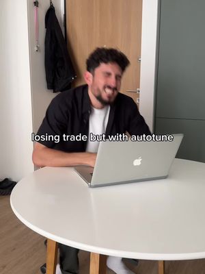 A man sits at a white table, smiling and looking at a laptop. He is in a room with a door and a jacket hanging on a wall. Text overlayed on the bottom says 'losing trade but with autotune'.