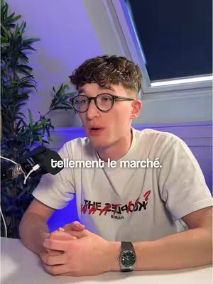 A young man wearing glasses and a white t-shirt is seated at a desk.  He has short, curly brown hair and fair skin. His shirt features text in a distressed, possibly reverse, style.  A microphone is visible to the left, and the background fades into purple and blue hues, illuminated by a window on the right.  The man wears a watch.  Appearing to speak directly to the camera.